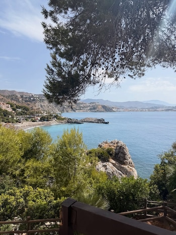 Vista aérea de una cala de arena bañada por aguas turquesas y rocas, rodeada de vegetación frondosa. Al fondo, colinas con edificios bajo un cielo azul claro