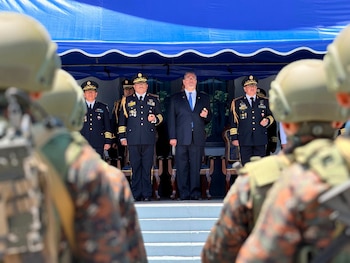Vista frontal de tres hombres uniformados con medallas y un hombre de traje, de pie bajo un toldo azul. En primer plano, personas con uniforme camuflado
