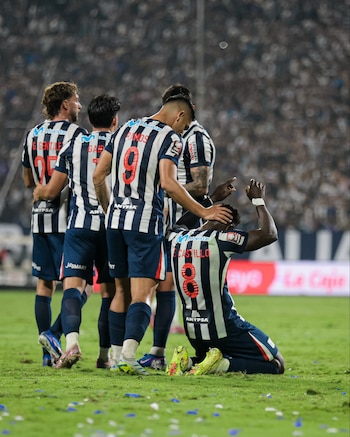 Varios jugadores de fútbol con camisetas a rayas blancas y negras celebran en un campo de césped verde. Un jugador arrodillado levanta los brazos y es felicitado