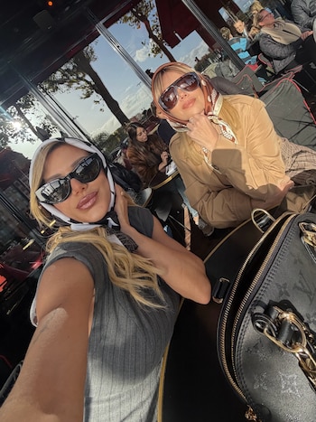 Dos mujeres con gafas de sol y pañuelos en la cabeza sentadas en una mesa al aire libre con un bolso en primer plano