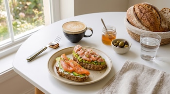 Plato con dos tostadas, una de aguacate y salmón troceado, otra de queso crema, aguacate y salmón ahumado. Café, miel, aceitunas, pan, vaso de agua, tenedor.