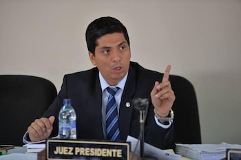 Primer plano de un hombre de mediana edad en traje oscuro y corbata azul a rayas, sentado en un escritorio con un cartel de "JUEZ PRESIDENTE", señalando con el dedo