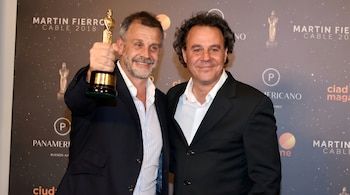 Dos hombres sonriendo a cámara, el de la izquierda levanta un trofeo dorado con base oscura. Llevan trajes oscuros. El fondo es un photocall con 'Martín Fierro Cable 2018'.
