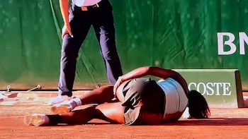 Una jugadora de tenis, Hailey Baptiste, está tumbada en el suelo de una cancha de arcilla roja, agarrándose la pierna, con un oficial de pie cerca