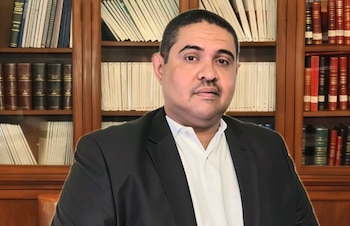 Hombre con bigote, traje negro y camisa blanca sentado frente a una estantería de madera llena de libros. El oficial mira directamente