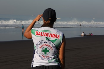 Vista trasera de una salvavidas con camiseta blanca y gorra azul, con la mano sobre la frente, mirando el océano y las olas en una playa de arena oscura