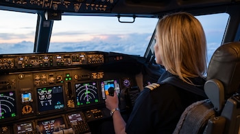 Una piloto de cabello rubio sentada de espaldas en la cabina de mando de un avión con pantallas iluminadas y el cielo de fondo.