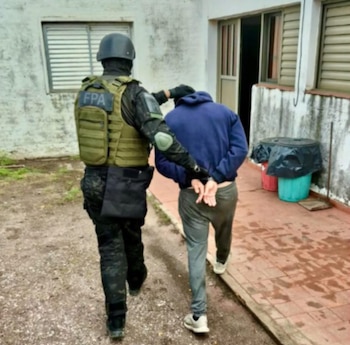 Un agente con chaleco táctico escolta por la espalda a un hombre esposado con buzo azul frente a una casa