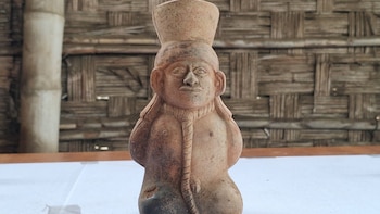 Escultura de cerámica antropomorfa de color marrón con una soga al cuello, colocada sobre una mesa frente a una pared tejida