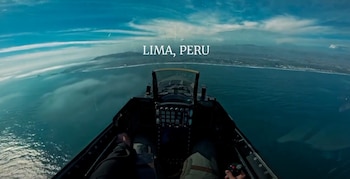 Vista desde la cabina de un F-16 mostrando el océano Pacífico y la costa de Lima, Perú, con nubes y el horizonte urbano al fondo. Se ven los controles del avión