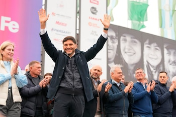 Maximiliano Pullaro, gobernador de Santa Fe, sonríe y saluda con los brazos en alto en un evento al aire libre, rodeado de gente que aplaude y una pantalla de fondo