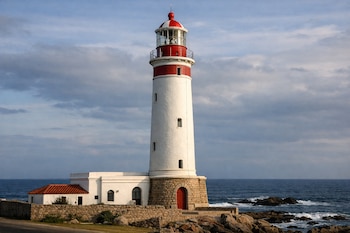 Faro cilíndrico blanco con bandas rojas y linterna, edificio blanco adyacente con tejado rojo, costa rocosa y el mar bajo un cielo con nubes.