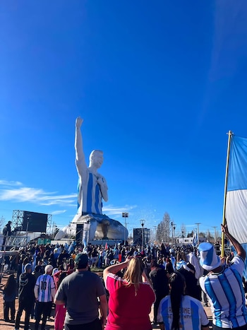Vista de una enorme estatua de Lionel Messi vestido con la camiseta argentina, con el brazo levantado, frente a una multitud de personas con banderas