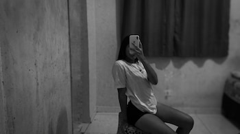 Fotografía en blanco y negro de una mujer sentada que oculta su rostro con un teléfono móvil blanco frente a una cortina.