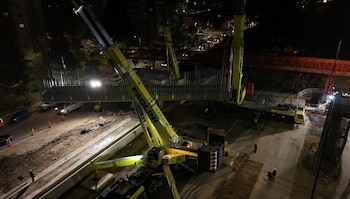 Grúas de construcción amarillas levantan una estructura metálica en un sitio de obra nocturno. Hay trabajadores y vehículos en la escena iluminada