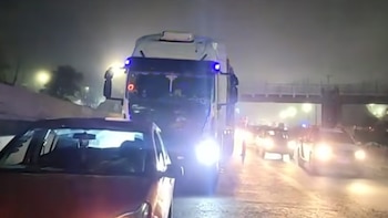 Camión blanco con luces azules y varios coches en una carretera con tráfico denso de noche. Hay neblina y los faros de los vehículos brillan