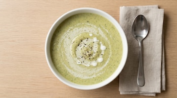 Plano cenital de un tazón blanco de sopa de crema de brócoli y manzana verde sobre madera clara, con servilleta de lino y cuchara metálica al lado.