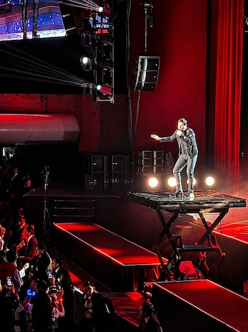 Chayanne canta sobre una plataforma elevadiza en un escenario con telones rojos, rodeado de luces, altavoces y una audiencia con teléfonos