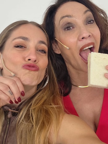 Dos mujeres. La de la izquierda con cabello rubio hace un gesto de beso. La de la derecha con cabello oscuro abre la boca y sostiene un sándwich. Ambas usan micrófonos