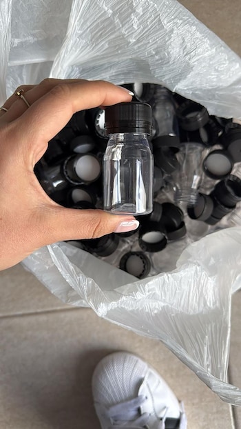 Una mano sostiene un pequeño envase transparente con tapa negra sobre una bolsa blanca llena de envases y tapas similares