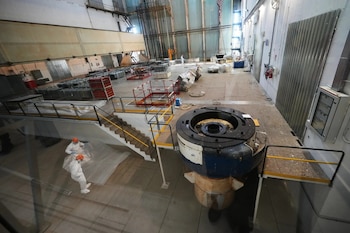 Interior de un gran espacio industrial. Dos personas con trajes blancos y cascos naranjas. Gran componente circular de maquinaria. Escaleras, barandillas, y equipos