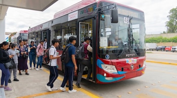 Una fila de estudiantes y jóvenes de diversas edades subiendo a un autobús rojo grande con la inscripción 'Binnibus' en el lateral, en una parada al aire libre.