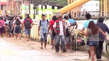 Un grupo de personas camina con pertenencias y bicicletas por una calle cubierta de agua y barro cerca de un puente