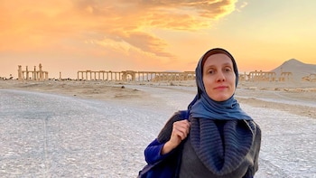 Una mujer, Shelly Renee Kittleson, con hijab azul y suéter oscuro, sonríe frente a las ruinas romanas de Palmira, Siria, bajo un cielo anaranjado al atardecer