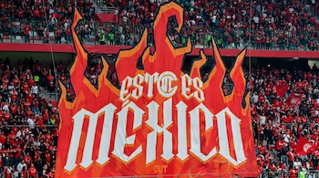 Pancarta roja y naranja con forma de llamas y la frase Esto es México en un estadio, con gradas llenas de aficionados y pequeñas banderas rojas.