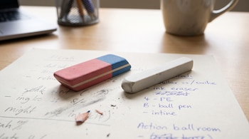Un borrador rosa y azul rectangular, un borrador blanco alargado, virutas de lápiz y una hoja de papel con texto manuscrito sobre un escritorio de madera.
