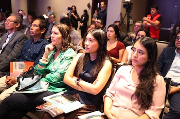 Personas sentadas en un auditorio escuchan una charla con folletos sobre las piernas y camarógrafos en el fondo