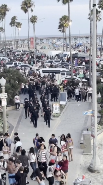 Agentes de policía caminan entre una multitud de personas en un paseo marítimo junto a una playa con palmeras, vehículos y un muelle al fondo