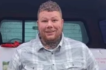 Un hombre de piel clara sonríe a la cámara, lleva una camisa a cuadros gris y blanca, y tiene tatuajes en el cuello. Fondo borroso