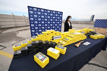 Mujer con mascarilla, mesa cubierta con tela oscura, paquetes amarillos y negros, letrero de 'EVIDENCIA', un rifle, carteles PDI, muro con alambre