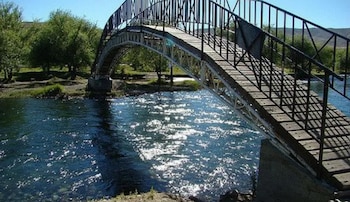 Un puente peatonal de madera y metal con estructura curva cruza un río de aguas claras rodeado de árboles