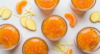 Vista cenital de varias gelatinas de mandarina y jengibre en vasitos transparentes, con rodajas y ralladura de jengibre, y gajos de mandarina.