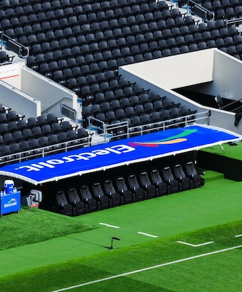Vista aérea de un estadio de fútbol con filas de butacas negras vacías. En primer plano, una banca azul con asientos cubiertos y parte de la cancha verde