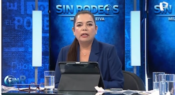 Milagros Leiva, presentadora de televisión, sentada en un set de noticias con fondo azul. Viste un blazer azul oscuro y tiene un tablet frente a ella