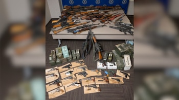 Una gran colección de fusiles, escopetas y pistolas, junto a cajas de municiones, exhibidos sobre una mesa y el suelo