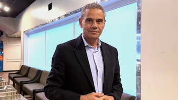 Hombre de mediana edad con traje oscuro y camisa clara de pie en una sala de espera con ventanas de tonos celestes y asientos marrones