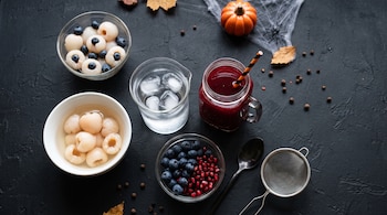 Cuencos con lichis y arándanos, vaso con hielo, jarra con jugo rojo, cuchara, colador y calabaza pequeña sobre una superficie negra.