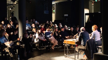 Una audiencia diversa sentada en sillas en un auditorio oscuro, mirando hacia dos oradores en una mesa con micrófonos bajo una luz brillante