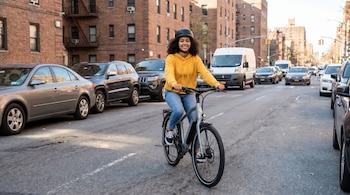 Adolescente con casco negro y sudadera amarilla pedalea una e-bike gris en una calle urbana con edificios de ladrillo y vehículos.