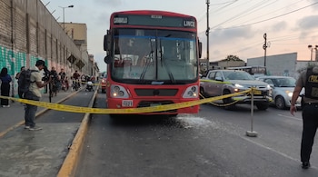 Autobús rojo detenido tras una cinta amarilla de seguridad, un policía a la derecha, un camarógrafo a la izquierda y vidrios sobre el asfalto.