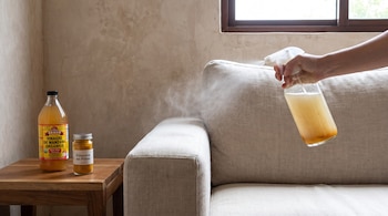 Una mano rocía un sofá con un atomizador. Sobre una mesa hay una botella de vinagre de manzana orgánico y un frasco de cúrcuma en polvo.