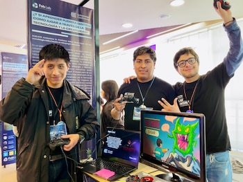 Tres jóvenes estudiantes sonríen mientras muestran un videojuego educativo en una exhibición, con dos sosteniendo controles y monitores con gráficos del juego