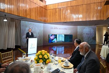 Hombre en traje habla en podio con logo "Banco de Guatemala", frente a pantalla con texto e imágenes. Personas sentadas en sillas, cortinas y flores visibles