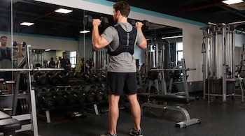 Hombre de espaldas con chaleco de peso, dos mancuernas, espejos, estantes de pesas y máquinas de ejercicio en un gimnasio.