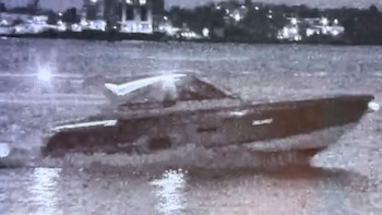 Vista nocturna de una lancha rápida oscura en el agua, con estelas claras detrás y luces difusas de una ciudad al fondo
