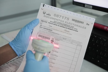 Mano enguantada de azul escaneando un documento electoral de ONPE con un lector de código de barras, mostrando parte del teclado de una laptop